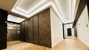 Un pasillo moderno con suelo de madera, un ascensor central y una larga galería con puertas a un lado.