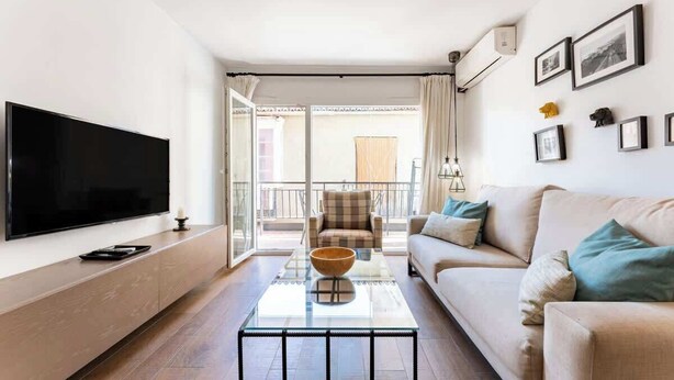 A modern living room with a flat-screen TV, a glass coffee table, a beige sofa, and a balcony with a view.