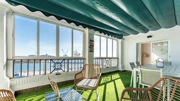 A sunlit room with a view of the sea, featuring a table and chairs, a small table with a vase, and a green artificial lawn.