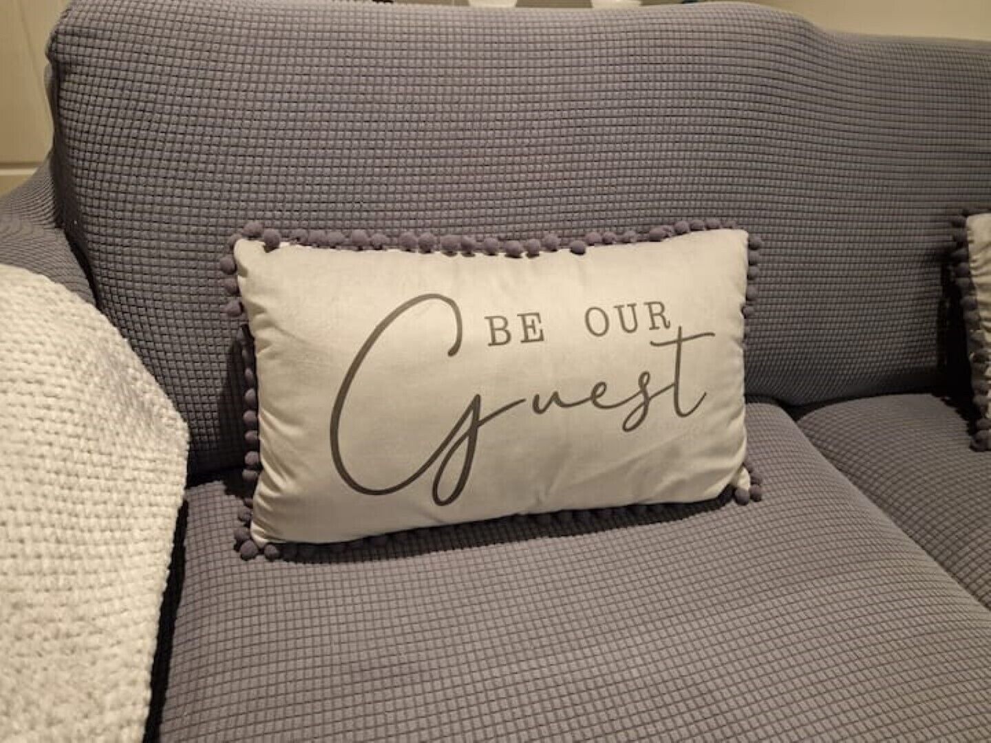 Un canapé gris avec un coussin sur lequel est écrit : « SOYEZ NOTRE INVITÉ ».