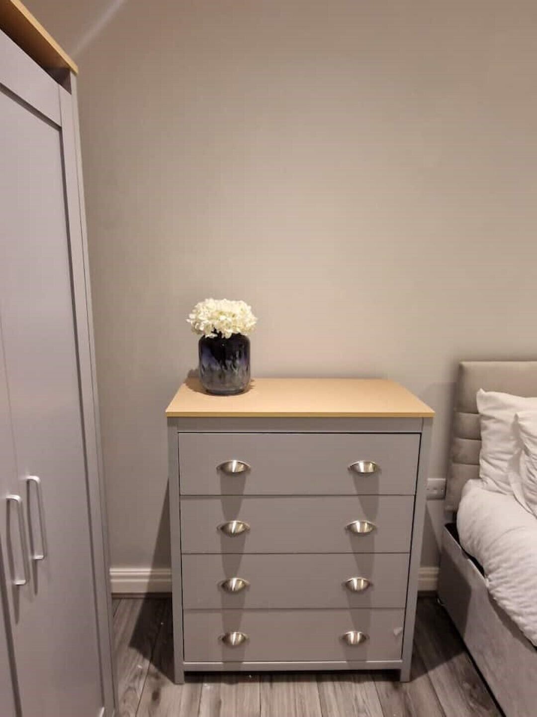 A bedroom with a bed, a grey nightstand with drawers, and a vase with flowers on the nightstand.
