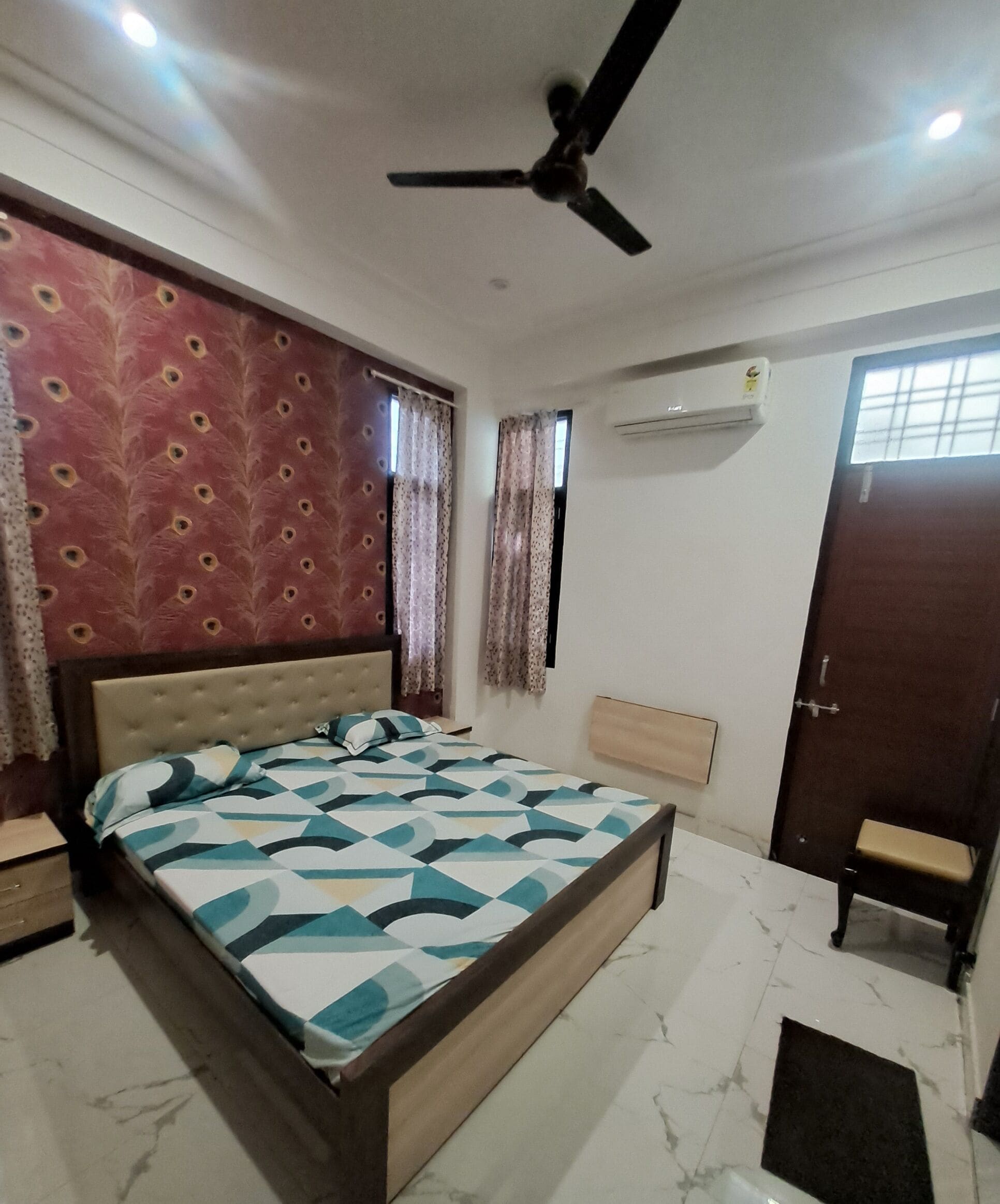 A bedroom with a bed, a ceiling fan, an air conditioning unit, a window with curtains, a door, and a bench.