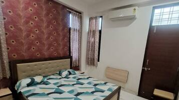 A bedroom with a bed, a ceiling fan, an air conditioning unit, a window with curtains, a door, and a bench.