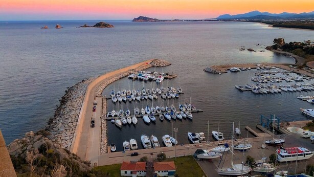 Un puerto deportivo con numerosas embarcaciones amarradas, una carretera que conduce al puerto y un paisaje montañoso al fondo.