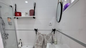 Une petite salle de bain comprenant des toilettes, une douche et un lavabo. Des étagères sont fixées au-dessus des toilettes pour y ranger les articles de toilette, et un miroir est accroché au mur.
