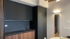 A modern kitchen with dark cabinets, a black countertop, and a wooden lower section. There is a sink with a faucet, a round table with a bowl, and a chair. - One bedroom apartment- Bandello 22 (Milan)
