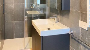 Une salle de bain moderne avec une douche à l’italienne en verre, un lavabo blanc et des murs recouverts de carreaux foncés.