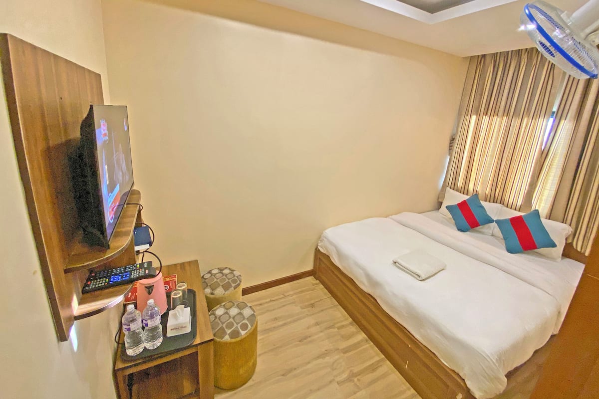 A hotel room with a bed, a TV mounted on the wall, a bedside table with a remote control, a water bottle, and a small lamp.