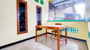 Una habitación con una mesa y silla de madera, una ventana con celosía metálica y una pared con adornos decorativos verdes.