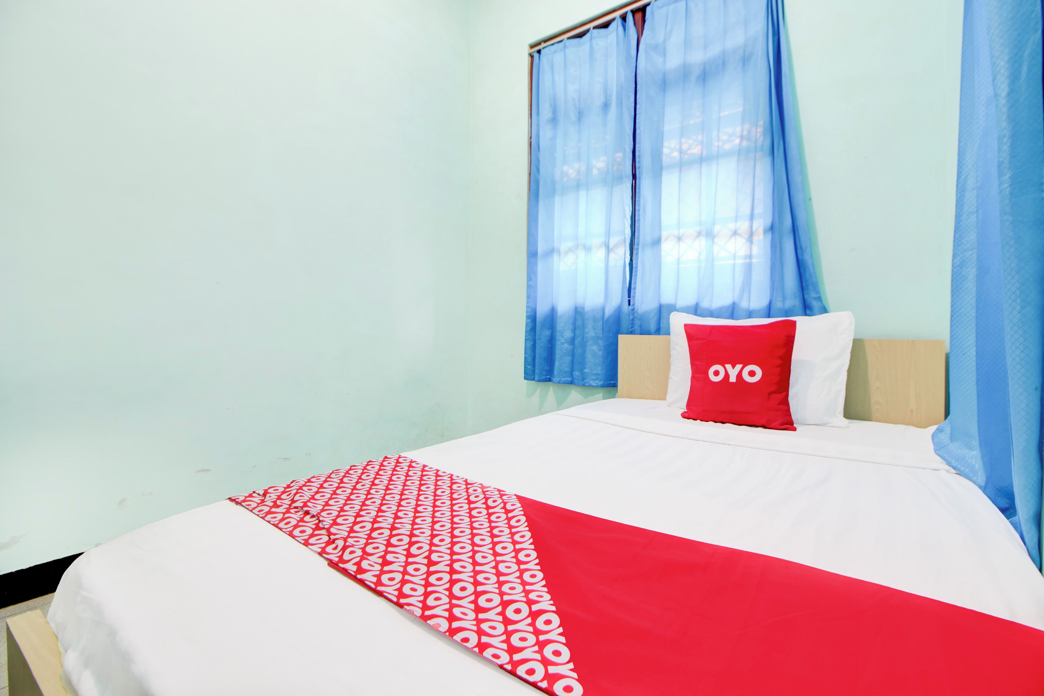 A neatly made bed with a red and white patterned blanket, a red pillow with the word "OYO" on it, and blue curtains on the window.