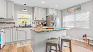 Une cuisine moderne avec des meubles blancs, un îlot central doté de tabourets, un réfrigérateur en acier inoxydable et une fenêtre avec des stores.