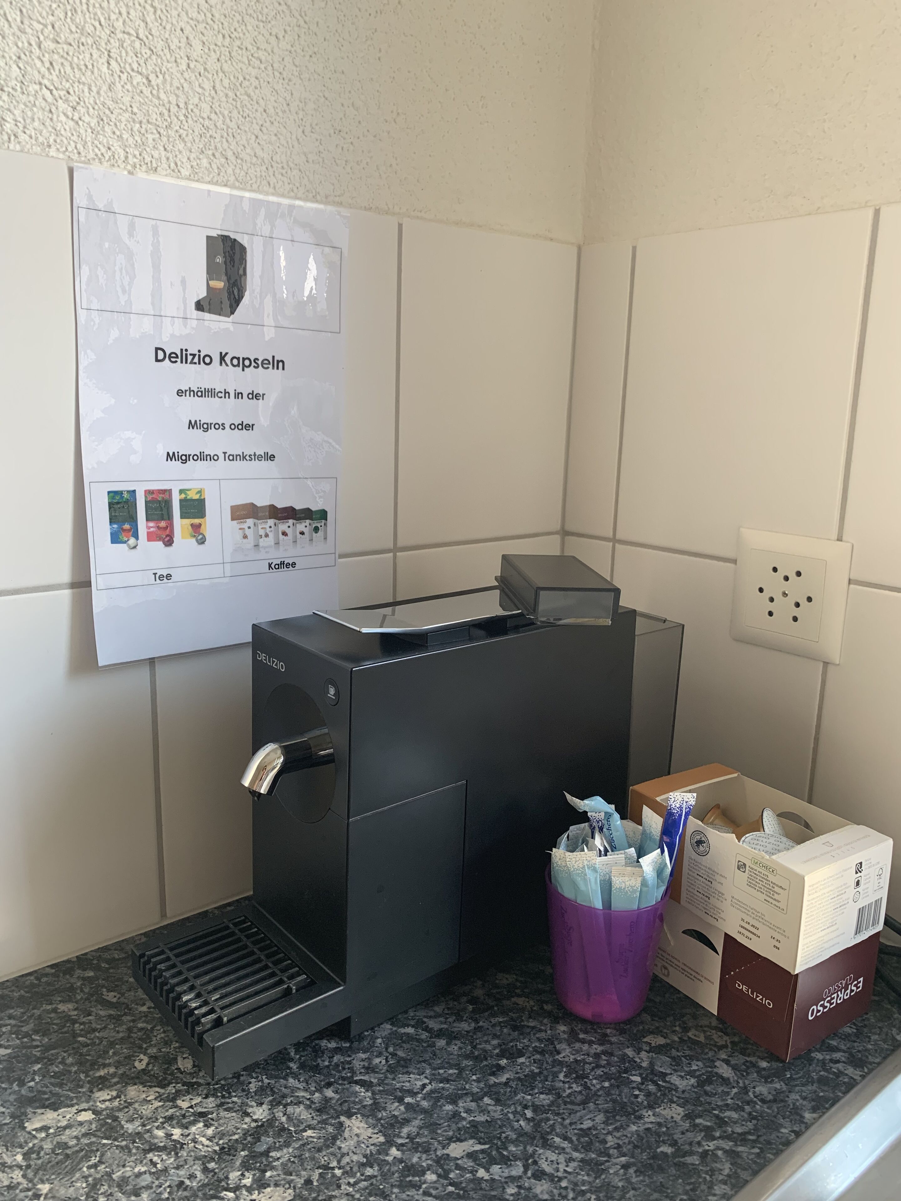 Una macchina per caffè con una vaschetta raccogligocce, un poster che pubblicizza capsule Delizio e una scatola di Espresso Delizio su un piano di lavoro in granito.