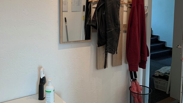 Een badkamer met een klok, een spiegel, een kapstok met jassen, een handdoekhouder, een desinfecterende handgeldispenser en een prullenbak.