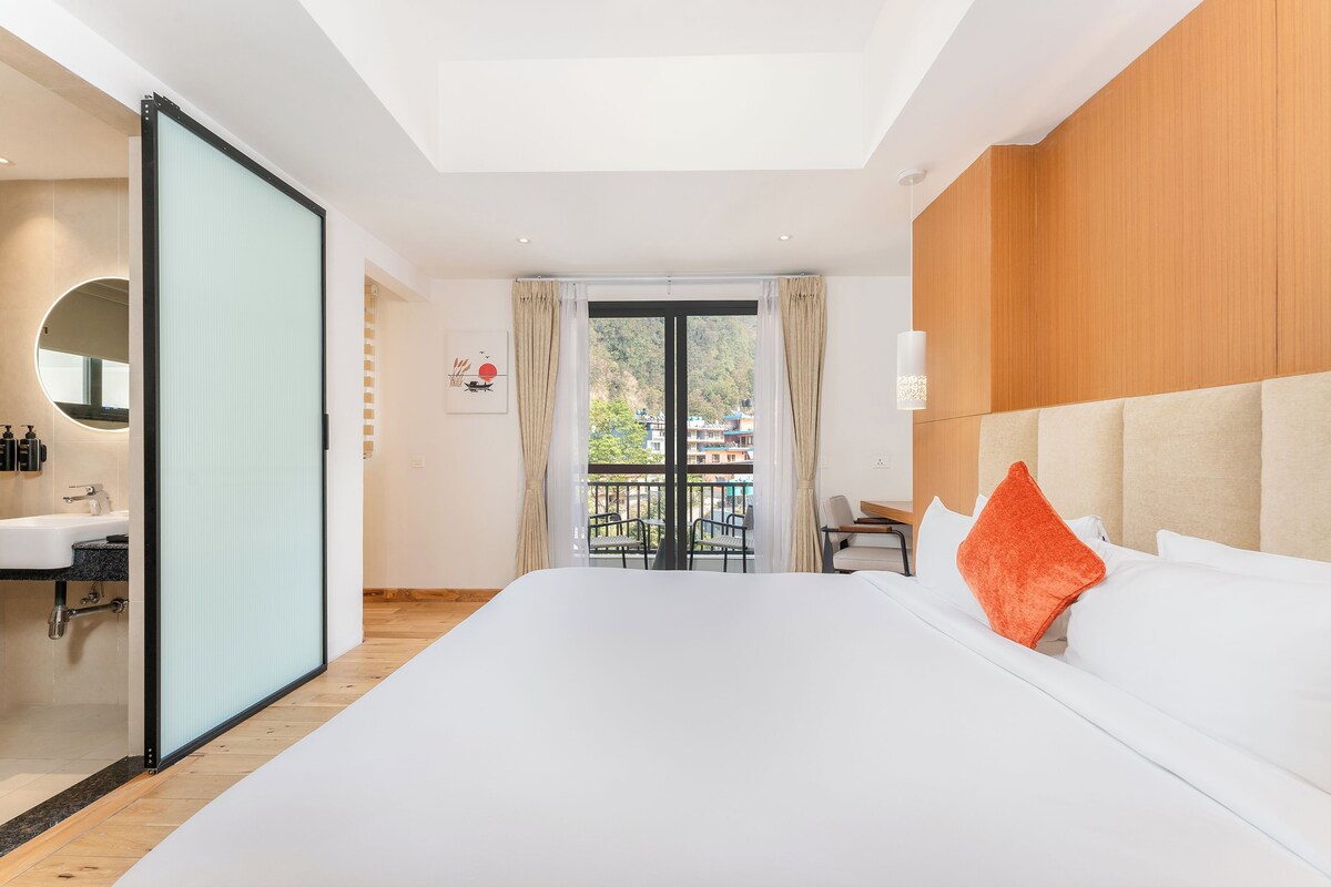 A hotel room with a large bed, a balcony with chairs, a desk, and a view of the outside.