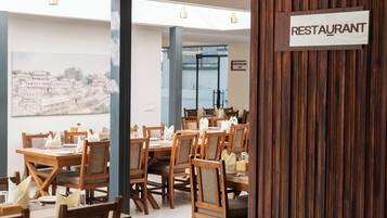 A restaurant interior with wooden tables and chairs, a sign indicating the restaurant, and a view of buildings through the windows.