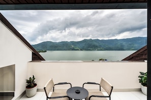 A balcony with a view of a lake and mountains, two wicker chairs, and two potted plants.