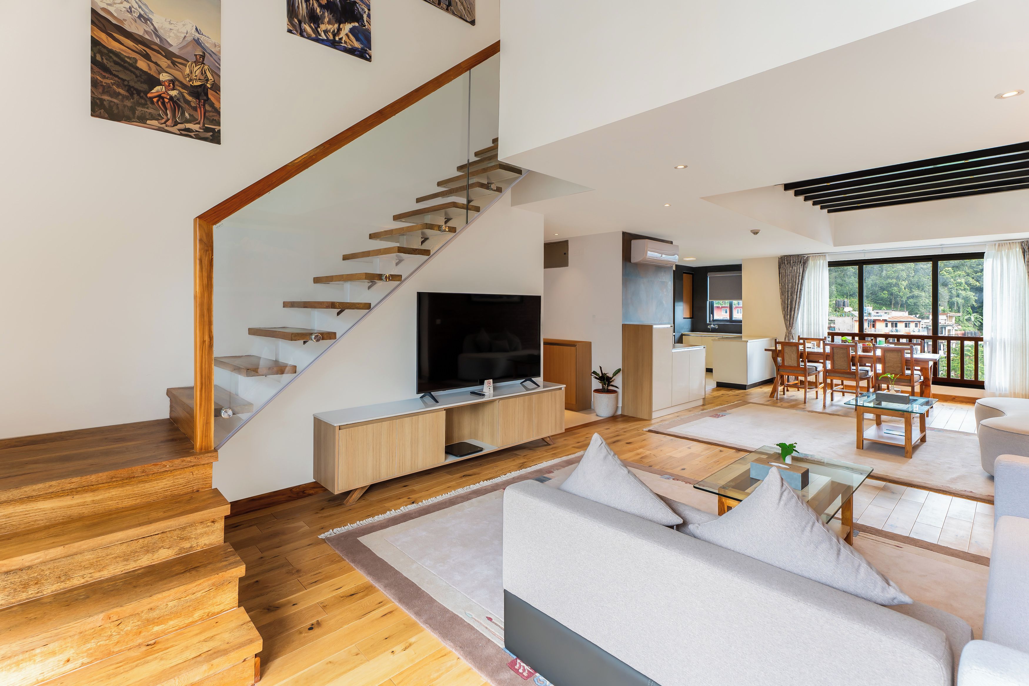 Una sala de estar moderna con una escalera de madera, un televisor de pantalla plana, un comedor con muebles de madera y un ventanal con vistas al exterior.