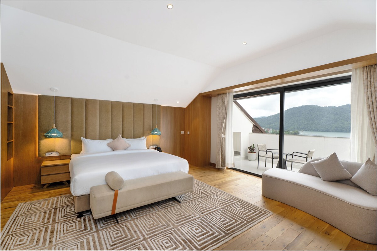 A modern hotel room with a large bed, a sofa, a balcony with a view, and a wooden floor.