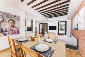 A dining area with a wooden table set for two, a white sofa, and a kitchen area with a TV and a stone wall. - Atithi Suites  (Pokhara)