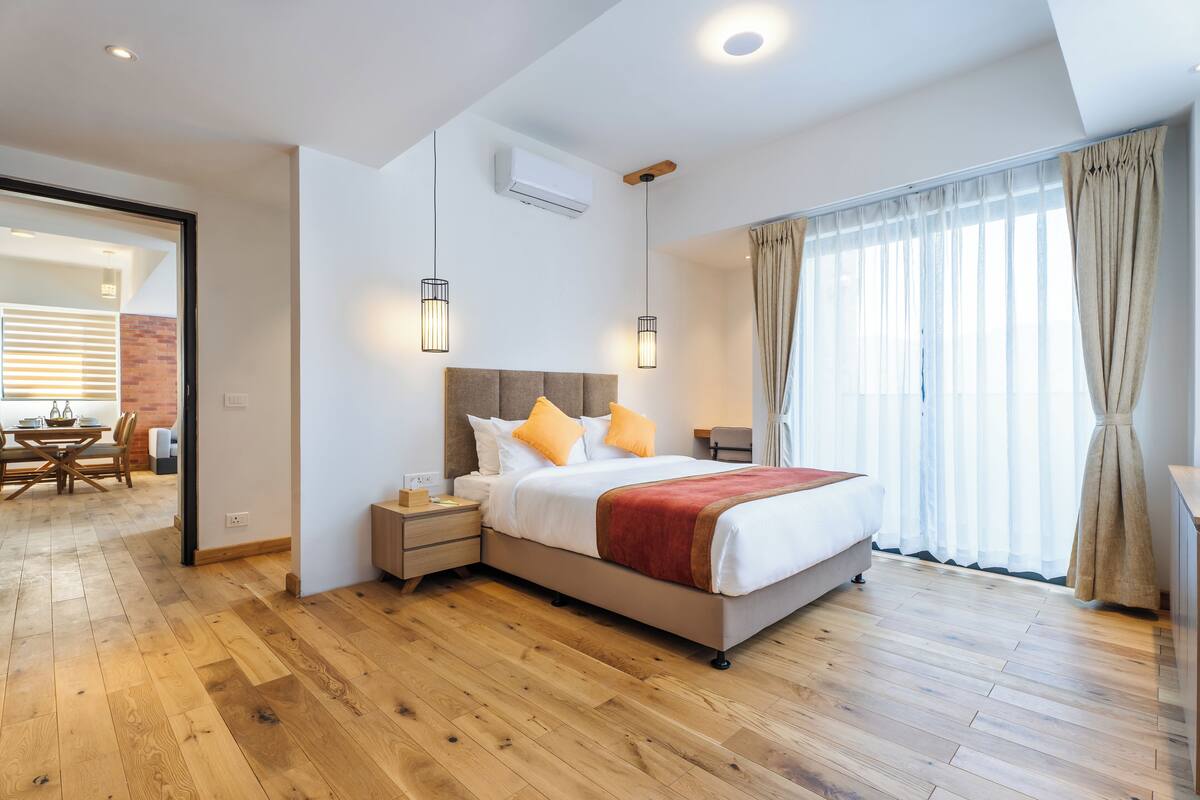 A modern bedroom with a wooden floor, a bed with white bedding and orange pillows, a nightstand, and a window with curtains.