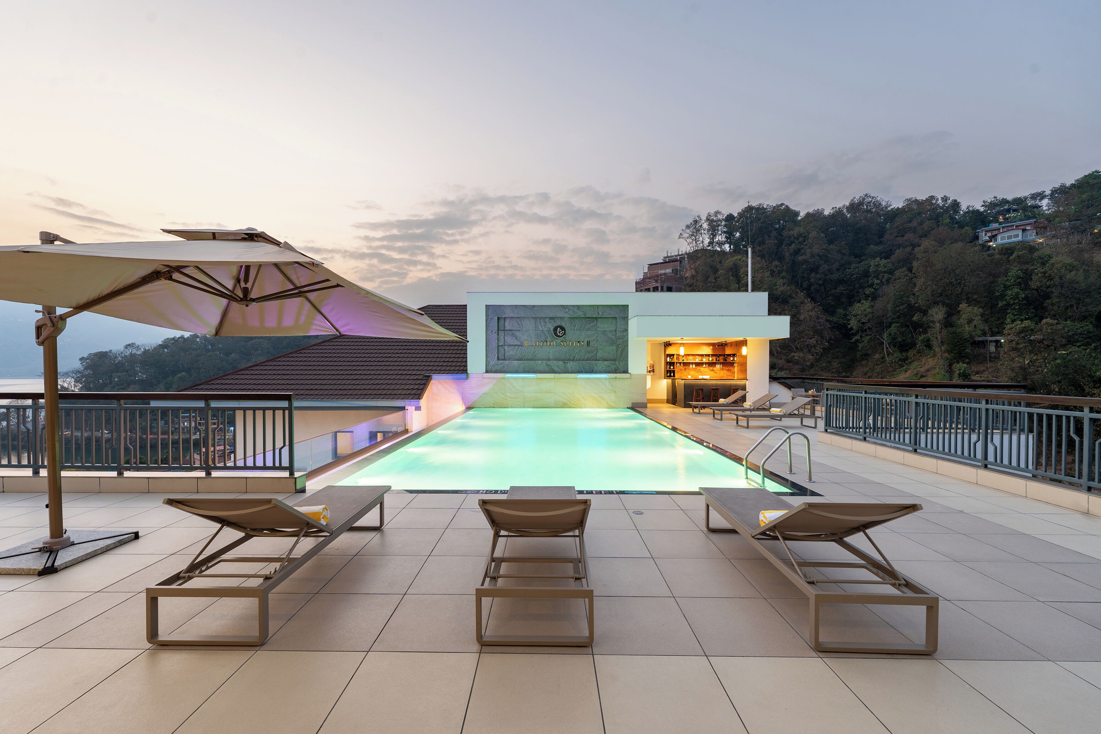 A rooftop pool area with lounge chairs and umbrellas, a bar, and a view of the surrounding landscape.