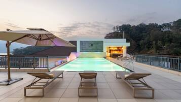 A rooftop pool area with lounge chairs and umbrellas, a bar, and a view of the surrounding landscape.