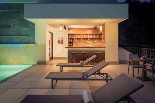 Un espace bar extérieur moderne, doté d’une piscine, de chaises longues et d’un comptoir bien garni de bouteilles.