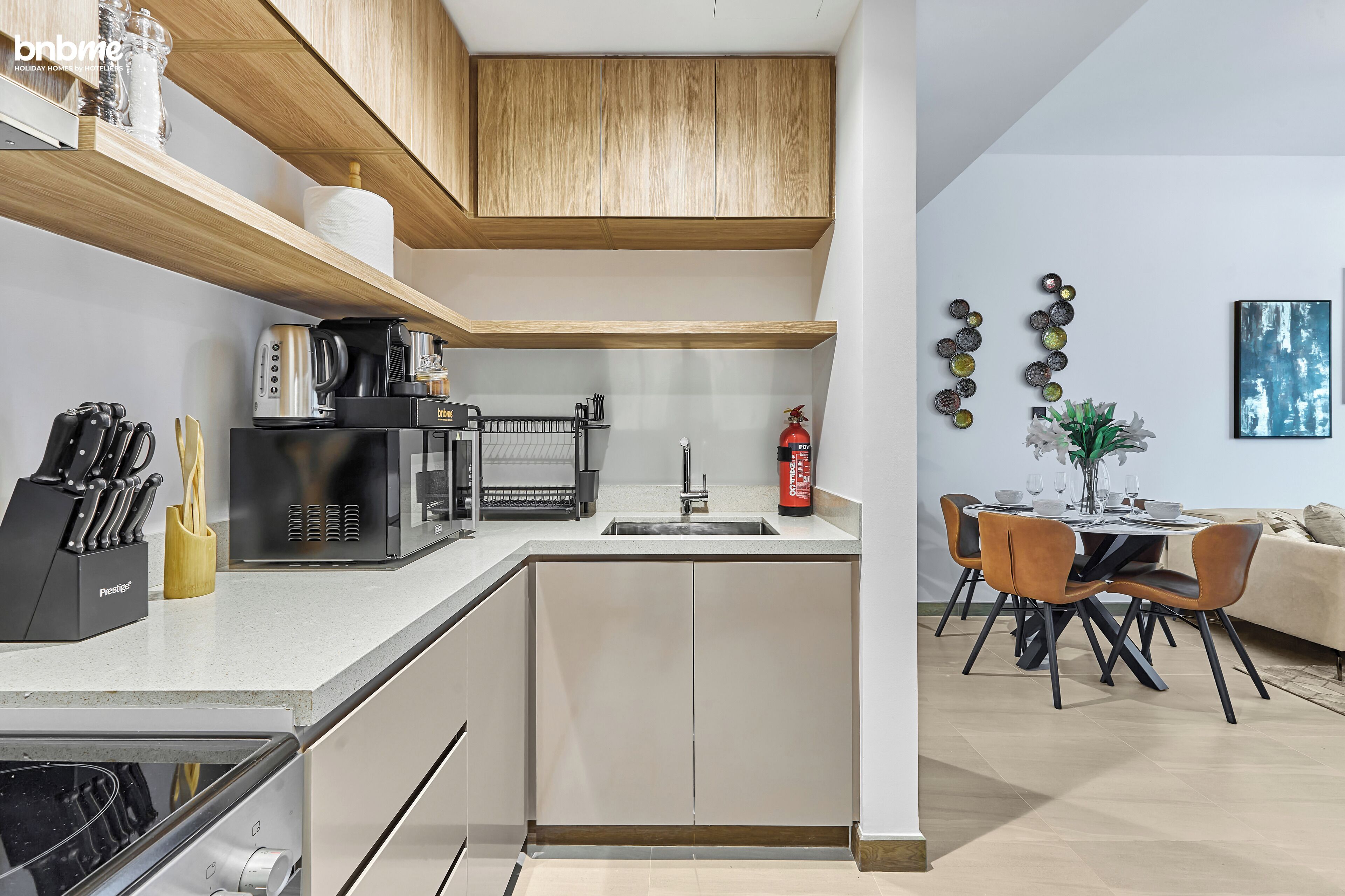 A modern kitchen with a built-in oven, microwave, and coffee maker. There is a sink with a faucet, and a fire extinguisher on the countertop. The kitchen has wooden cabinets and a white countertop.