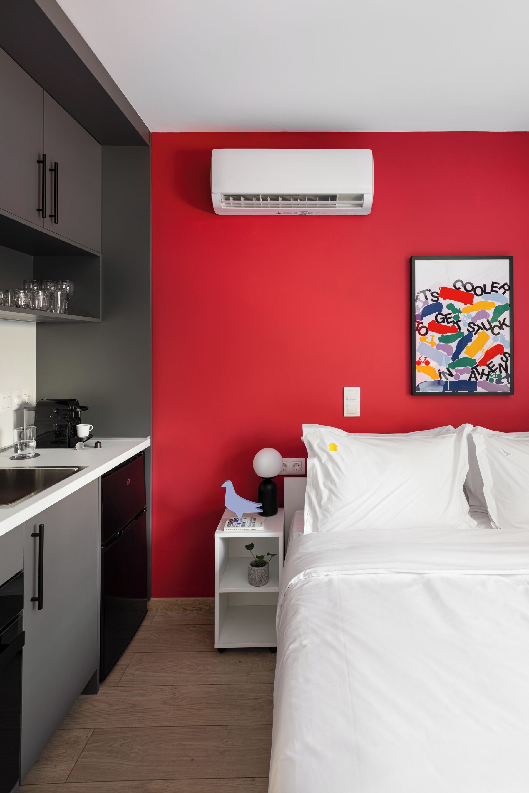 A compact room with a kitchenette, a bed with white bedding, a red accent wall, and a colorful artwork.