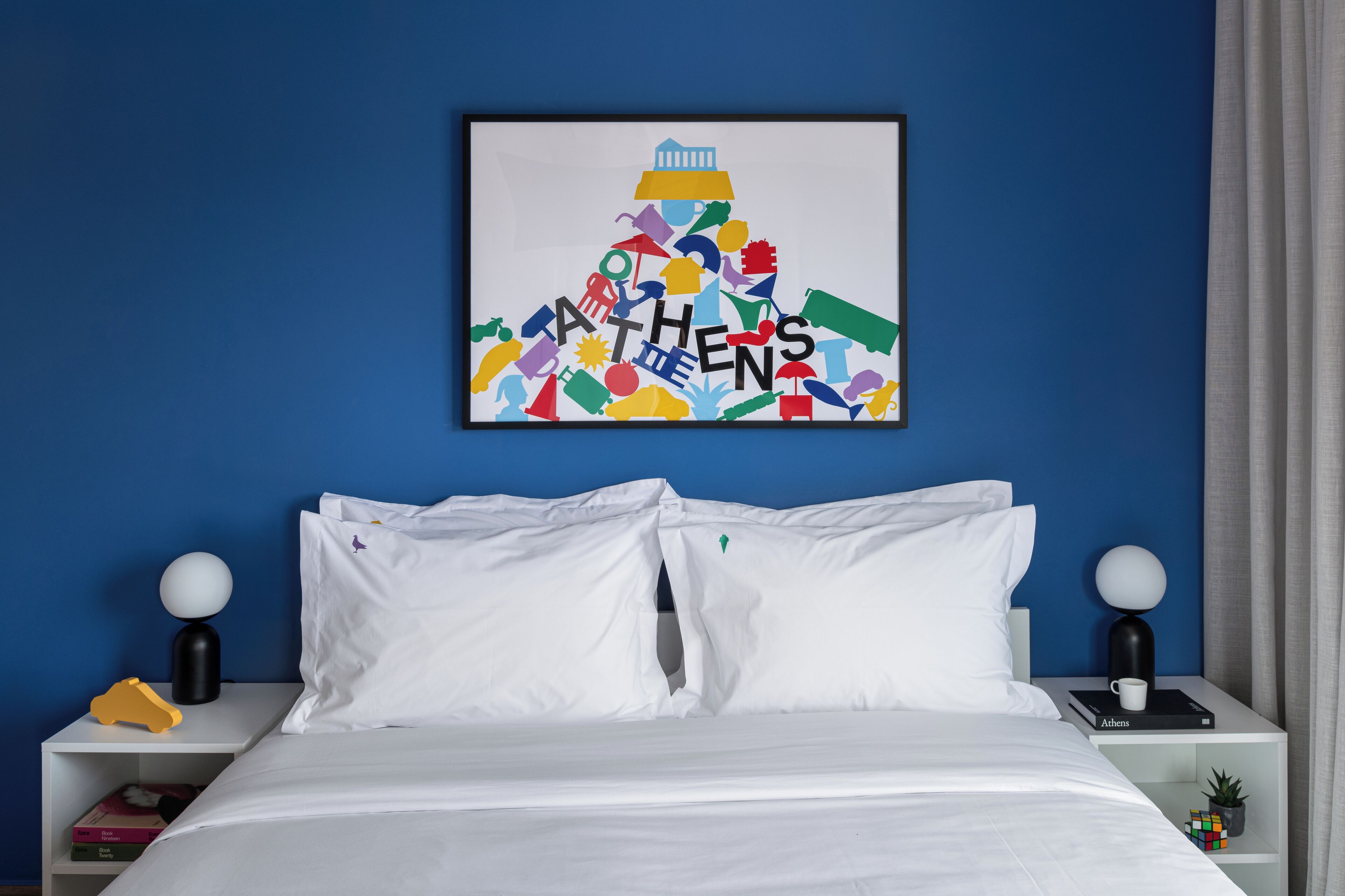 Una habitación de hotel moderna con una pared azul de acento, una cama blanca con dos almohadas, una obra enmarcada que representa un paisaje urbano colorido y una mesita de noche con una lámpara y un libro titulado 'Atenas'.