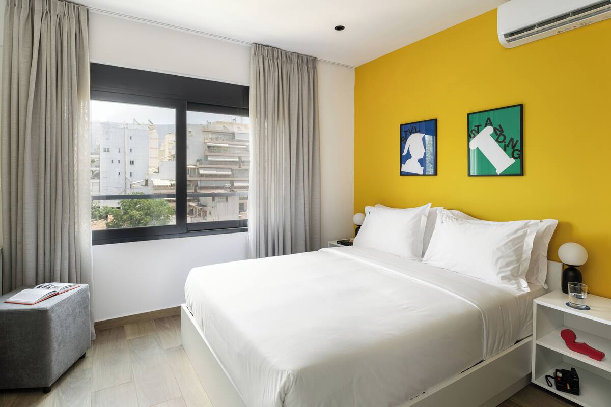 Ein Hotelzimmer mit einem großen Bett, einem Nachttisch mit Lampe, einem kleinen Hocker mit einem Buch und einem Fenster mit Vorhängen.
