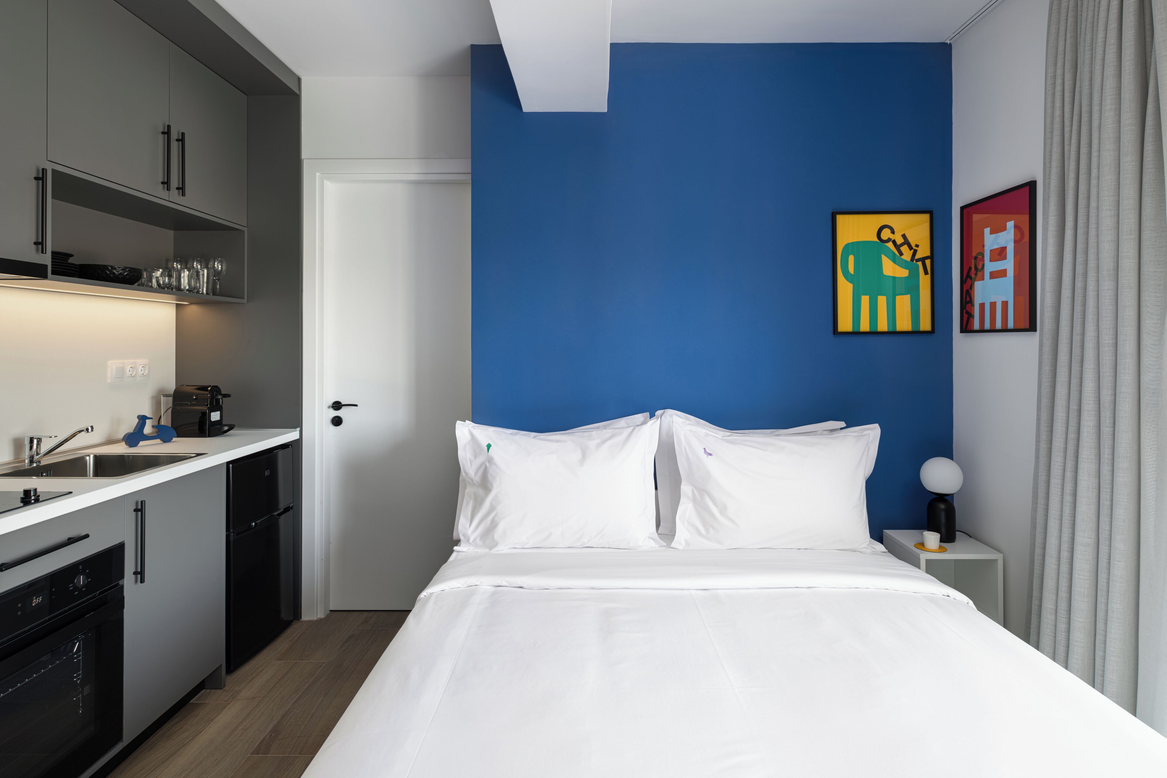 Una habitación de hotel moderna con una cama blanca, un muro azul decorativo y una zona de cocina con armarios grises y una encimera blanca.