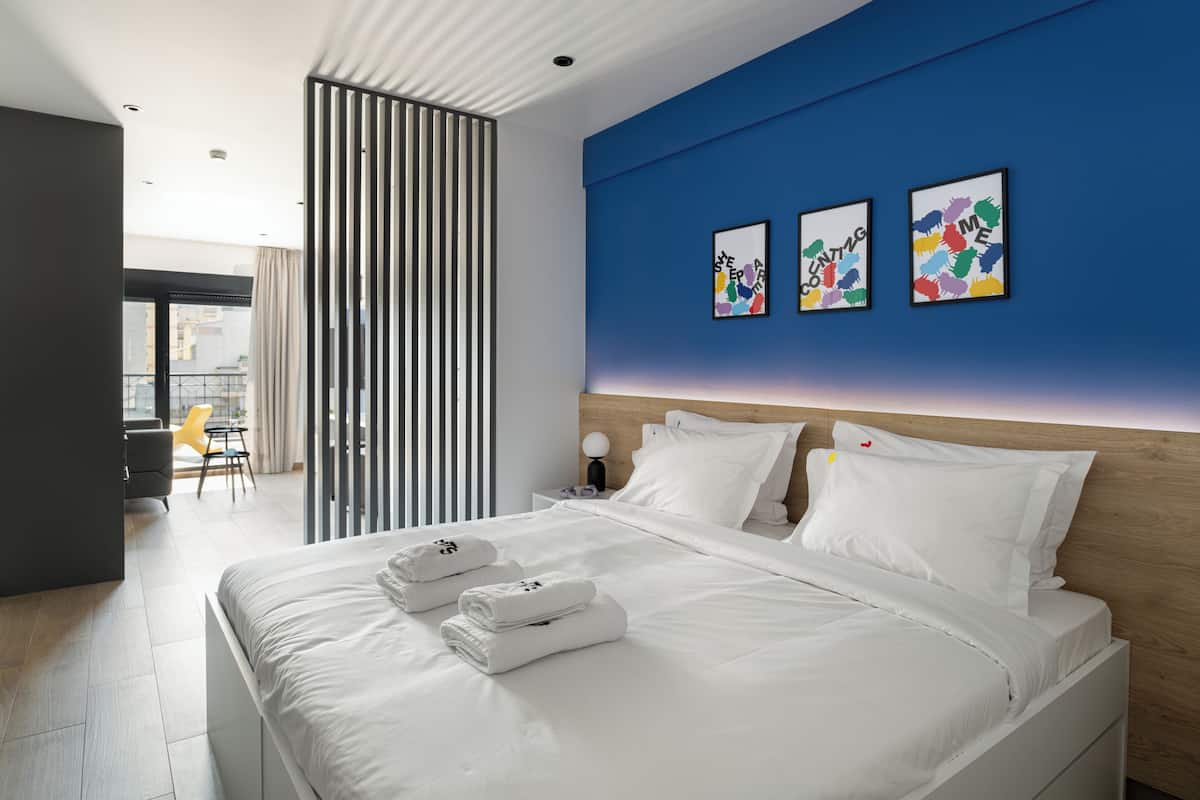 Ein modernes Hotelzimmer mit einem großen Bett, weißer Bettwäsche und einer blauen Akzentwand. Über dem Bett hängen drei gerahmte Kunstwerke, und durch das Fenster ist die Stadt zu sehen.