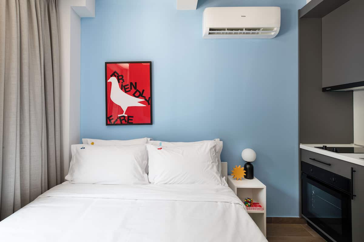 Ein Hotelzimmer mit einem Bett, weißen Kissen, einem gerahmten Poster, einem Nachttisch mit Lampe und einer Küchenzeile mit Herd.