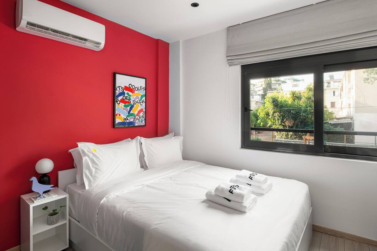 Ein Hotelzimmer mit einem Bett, weißer Bettwäsche, einer roten Akzentwand, einem Fenster mit Blick auf Grünflächen und einer Wand-Klimaanlage.