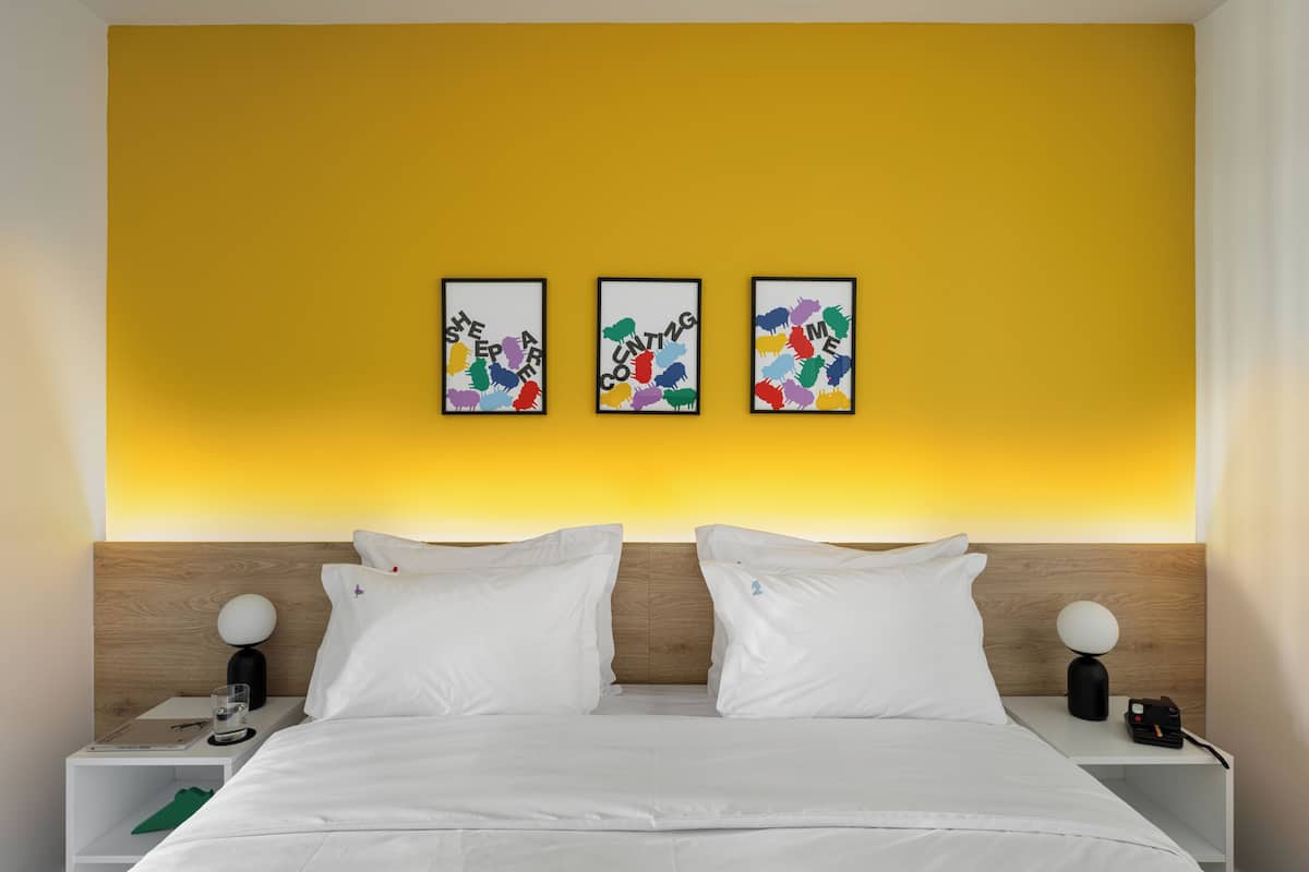 Ein Hotelzimmer mit einem Bett, weißer Bettwäsche, einem hölzernen Kopfteil, zwei Nachttischen mit Lampen und drei gerahmten Kunstwerken an der Wand.