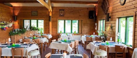 A cozy wooden cabin-style restaurant with multiple tables set for dining, featuring natural light from large windows and a rustic interior.