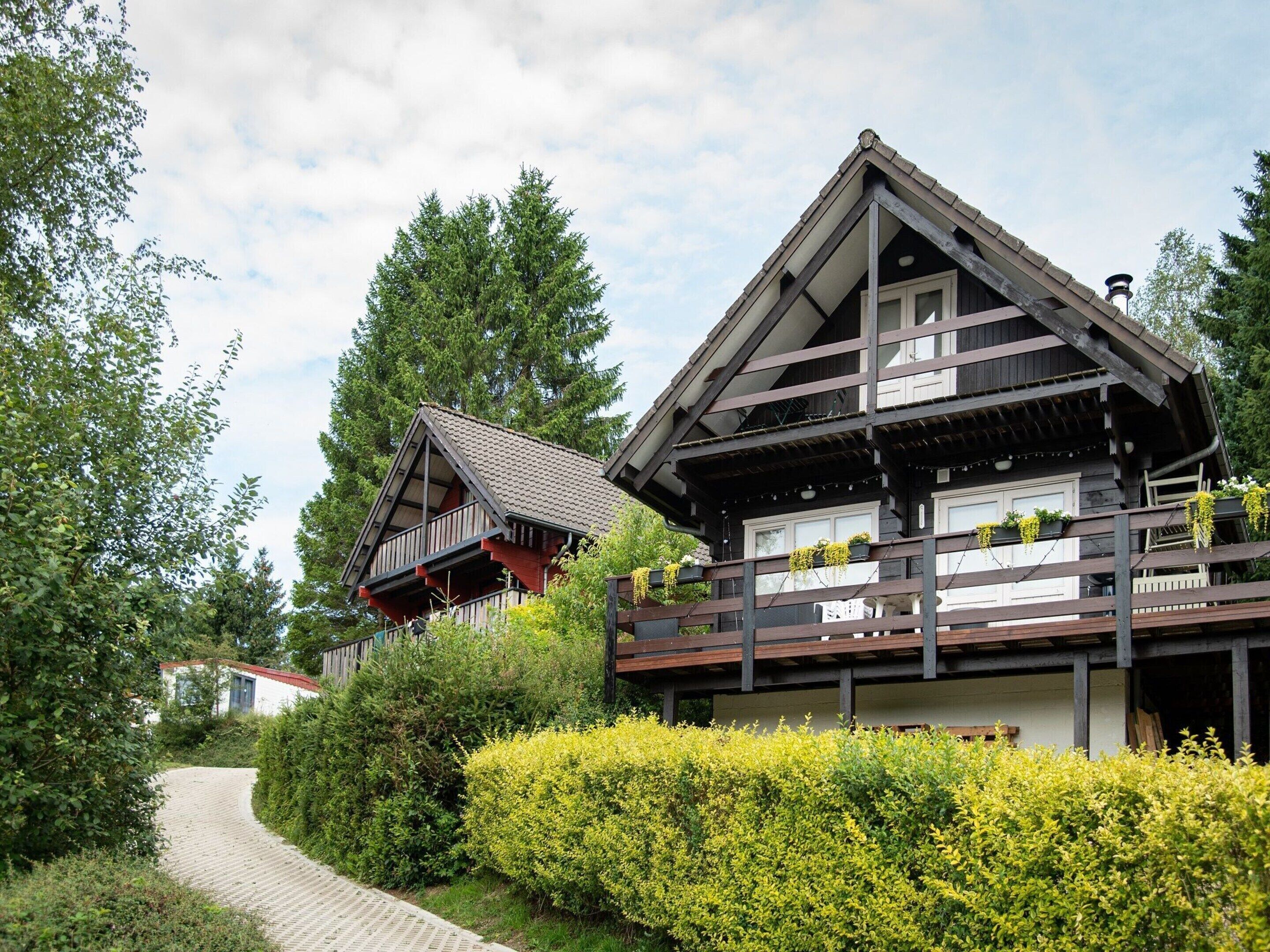 A traditional chalet with a wooden balcony and a sloped roof, surrounded by lush greenery and a paved path.