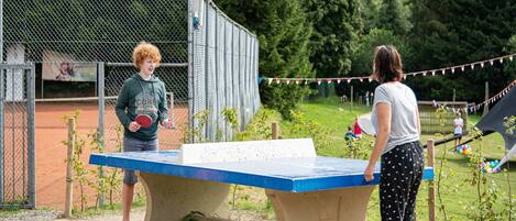 A table tennis table in an outdoor setting with a chain-link fence and greenery in the background.