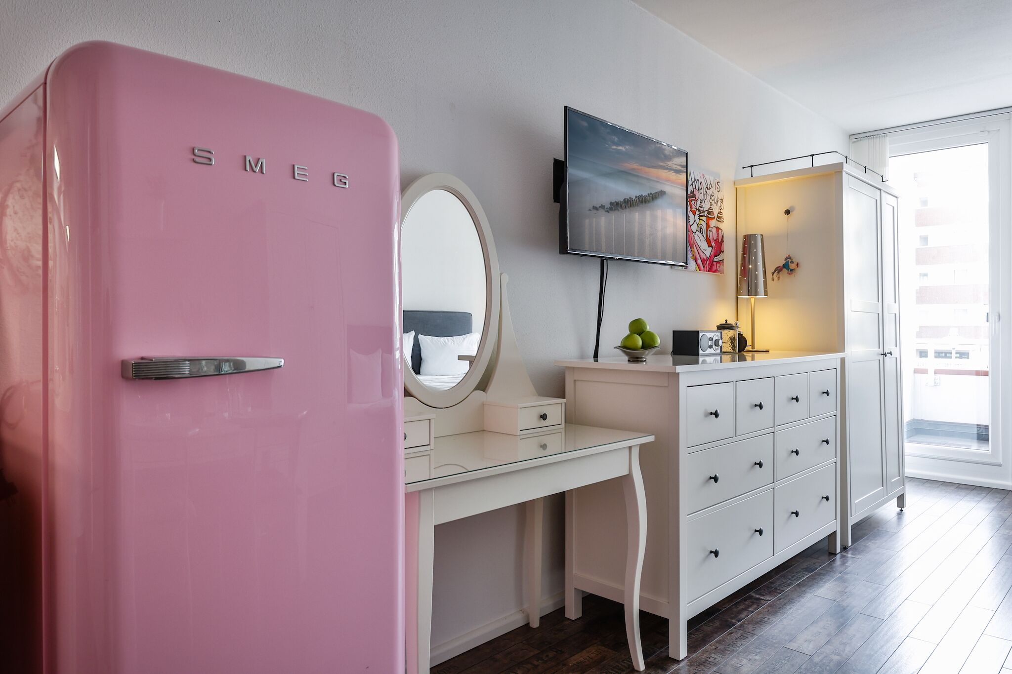 Een roze koelkast, een wit bureau met lades, een aan de muur bevestigde televisie en een raam met uitzicht op gebouwen.