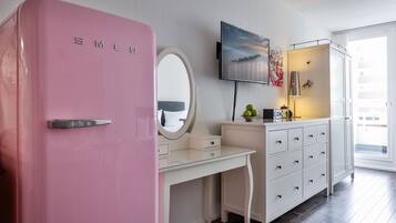 Een roze koelkast, een wit bureau met lades, een aan de muur bevestigde televisie en een raam met uitzicht op gebouwen.