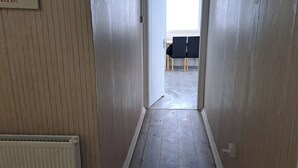Ένας διάδρομος με ξύλινο δάπεδο και λευκούς τοίχους, που οδηγεί σε ένα δωμάτιο με ένα τραπέζι και καρέκλες.
