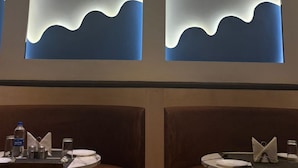 Restaurante com três janelas iluminadas exibindo um padrão de ondas. Mesas montadas com pratos, talheres e garrafas de água.