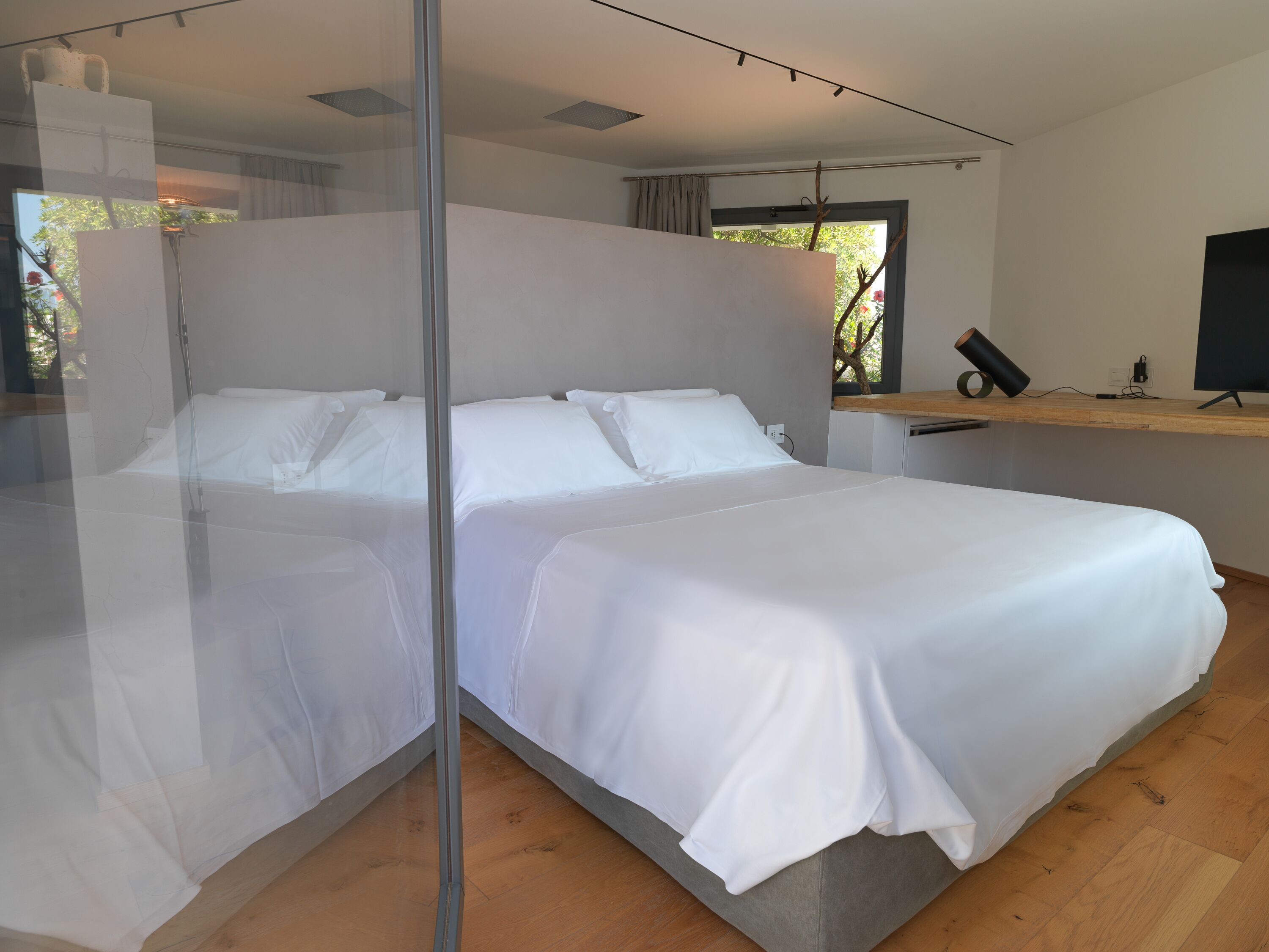 A modern bedroom with a large bed, a bedside table, a mirror, and a window with a view of trees.