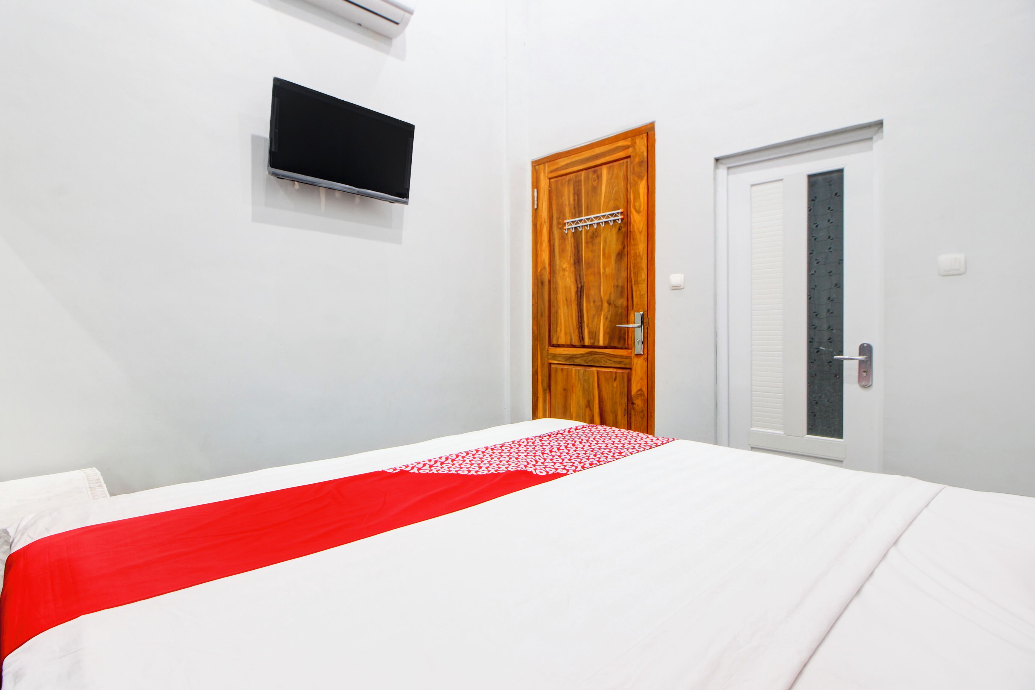 Une chambre à coucher avec un lit, une porte en bois, un mur blanc et un téléviseur fixé au mur.