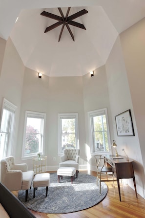 A spacious room with a high ceiling, large windows, a round rug, and a desk with a chair.