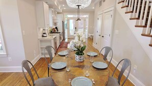 A dining area with a wooden table set for six, surrounded by chairs, a chandelier, and a view to the kitchen.