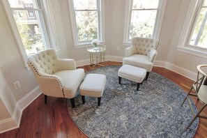 A room with two white tufted armchairs, a matching ottoman, a round blue rug, and a small glass table with a vase.