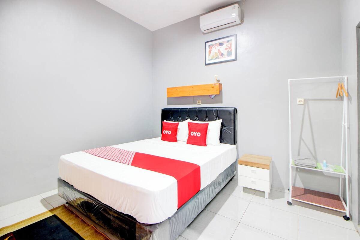 Habitación de hotel impecable con una cama grande, una mesita de noche, un pequeño tocador de madera, un armario blanco y un estante fijado a la pared.