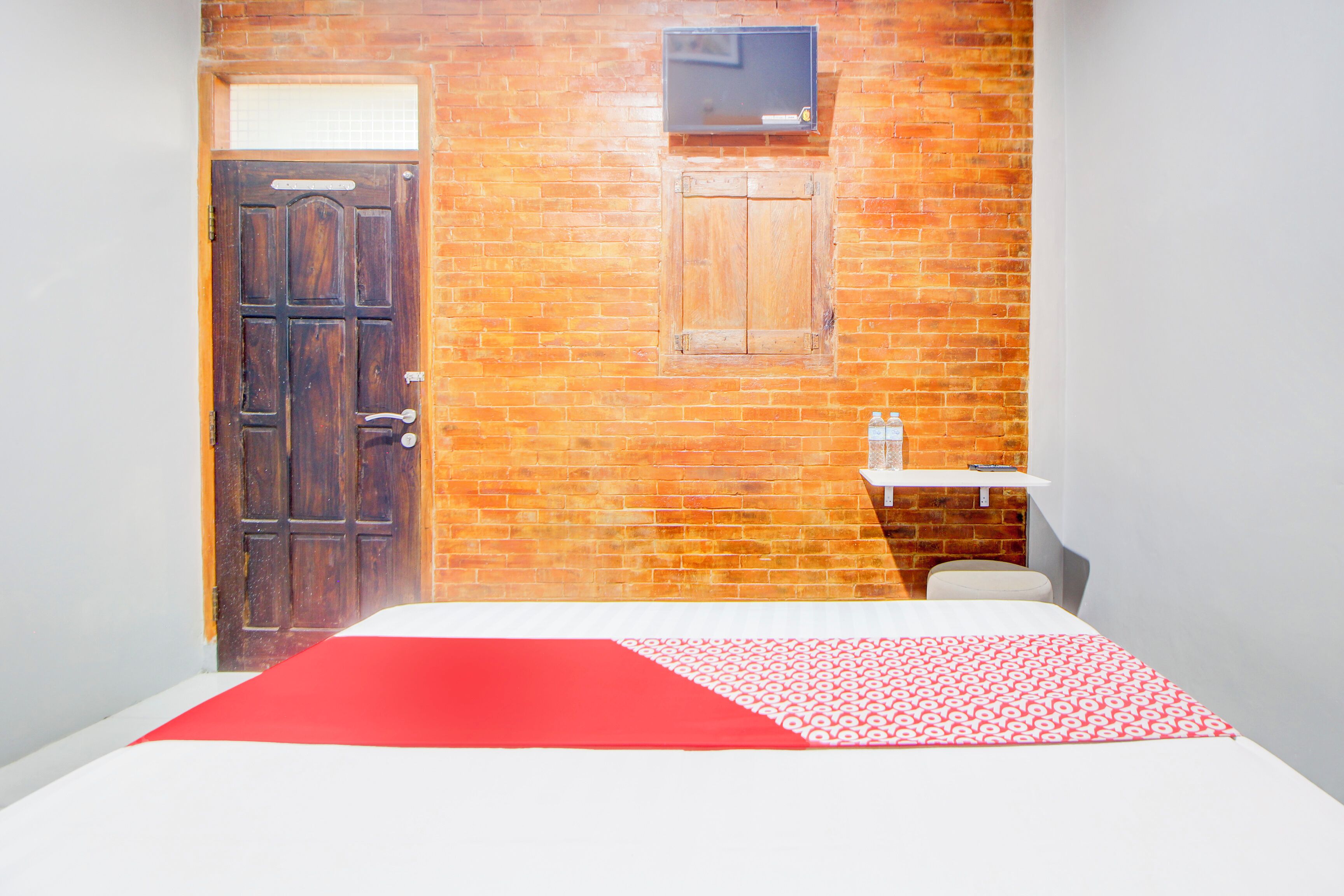 Um quarto com parede de tijolos, porta de madeira, uma prateleira pequena com uma garrafa e uma cama com colcha de padrão vermelho e branco.
